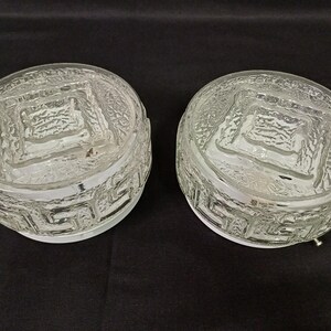 May include: Two clear glass light fixtures with a textured surface and a geometric design around the base. The fixtures are round and have a square pattern in the center. The glass is clear, and the fixtures are on a dark background.