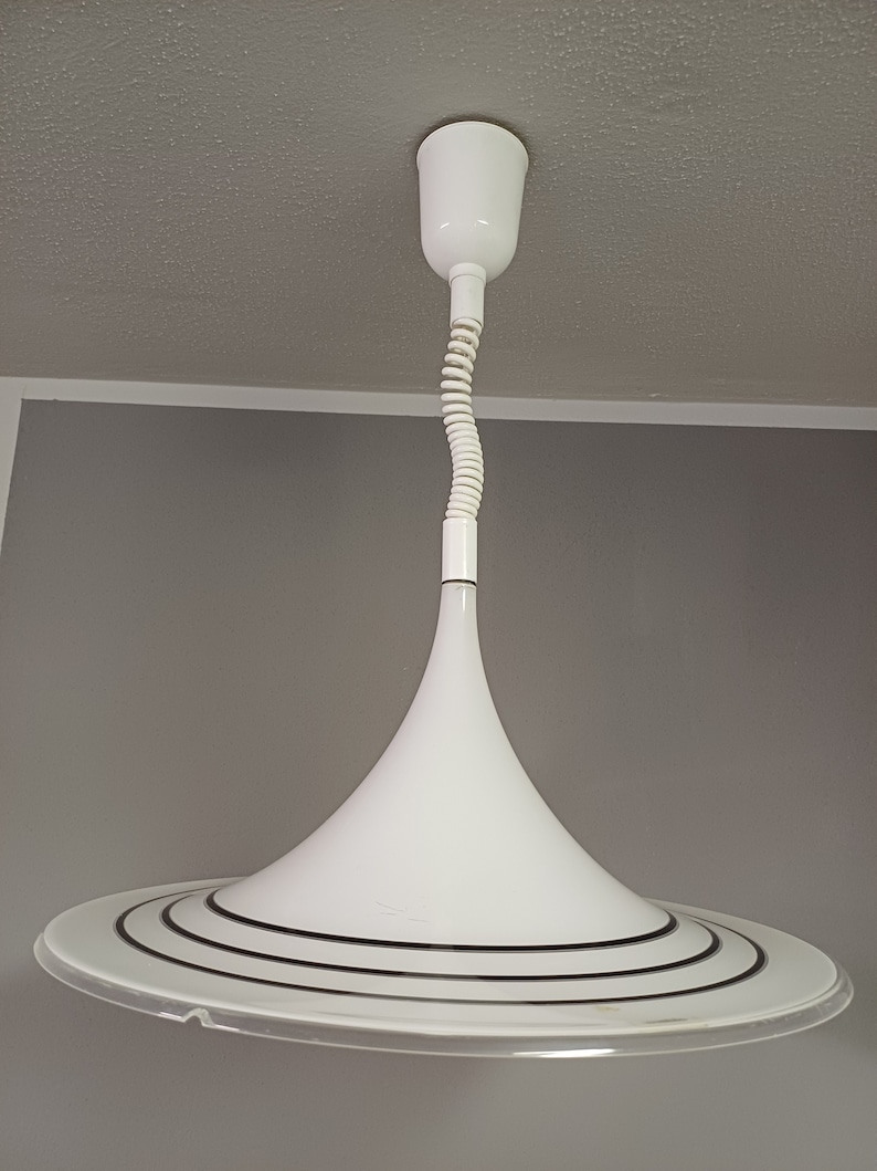 May include: White pendant light with a white, coiled cord and a white, cone-shaped shade with a black, concentric ring design.