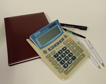 May include: A beige calculator with blue accents, a maroon notebook, two white pens, and two black markers. The calculator has a digital display and a numeric keypad. The notebook is closed and has a stitched border.