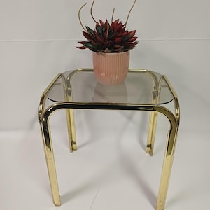 Może przedstawiać: Stolik pomocniczy w stylu vintage ze złotą metalową ramą i przezroczystym szklanym blatem. Mały, różowy, żebrowany ceramiczny garnek zawiera roślinę z czerwonymi i zielonymi liśćmi i smukłymi, zakrzywionymi łodygami. Stół ma kwadratowy kształt.