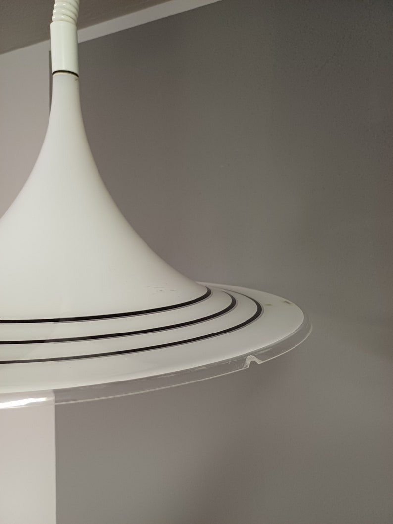 May include: White pendant light with a wide, circular base and a narrow, cone-shaped shade. The base has three black rings around the edge.