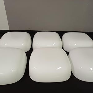 Könnte beinhalten: Sechs weiße, abgerundete quadratische Objekte sind auf einer schwarzen Oberfläche angeordnet. Die Objekte haben eine glänzende Oberfläche und scheinen aus einem festen Material zu bestehen. Die Beleuchtung erzeugt Glanzlichter auf den gekrümmten Oberflächen.