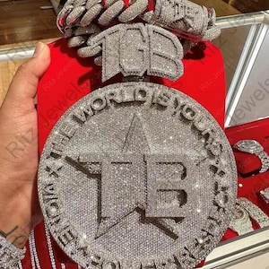 May include: A diamond-encrusted pendant with the text "THE WORLD IS YOURS" and the initials "TB". The pendant is round with a star in the center. A matching diamond chain bracelet is also visible.