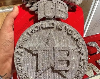 May include: A diamond-encrusted pendant with the text "THE WORLD IS YOURS" and the initials "TB". The pendant is round with a star in the center. A matching diamond chain bracelet is also visible.