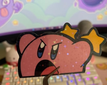 May include: A pink and yellow holographic sticker of a cartoon character with a crown. The character has a pink face, blue eyes, and a wide open mouth. The sticker is on a white keyboard.