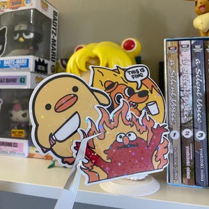 May include: A sticker featuring a cartoon character with red fur and a yellow beak, engulfed in flames. The character is holding a cup of coffee and has a surprised expression. The sticker has a holographic finish and the text "This is fine" is written in a speech bubble above the character.