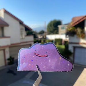 May include: A purple and iridescent sticker of a cartoon character with a smiling face and a star pattern. The sticker is held by tweezers.