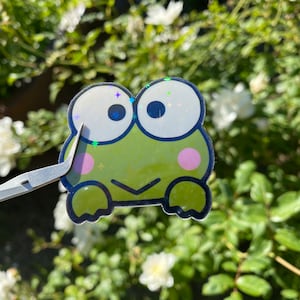 May include: A green frog sticker with large, white eyes and pink cheeks. The sticker has a holographic finish and is surrounded by a black outline.