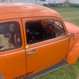 May include: A bright orange Volkswagen Beetle with a black "Jägermeister" sticker on the side. The car has a yellow smiley face sticker on the door.