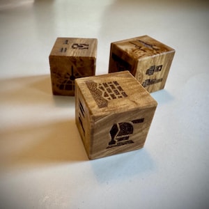 May include: Three wooden dice with etched designs. Each die features a different icon, including a clock, a telephone, and a building. The dice are made of light-colored wood and have a natural grain pattern. The dice are arranged on a white surface.