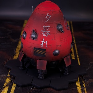 May include: A red, egg-shaped model with a hexagonal base. The model has multiple small, round details and a hatch on the side. The model has Japanese characters written on the side: "分身機".