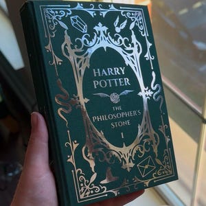 May include: A green hardcover book with silver foil detailing. The title is "Harry Potter and the Philosopher's Stone" with the number "1" below. The book is held in a hand.