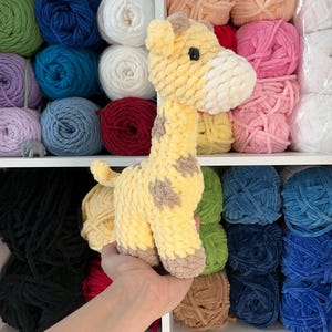 Pu&ograve; includere: Una giraffa all'uncinetto gialla e marrone con macchie marroni. La giraffa &egrave; tenuta davanti a una mensola piena di gomitoli di lana di vari colori, tra cui blu, verde, rosa e bianco. La giraffa &egrave; un giocattolo morbido.