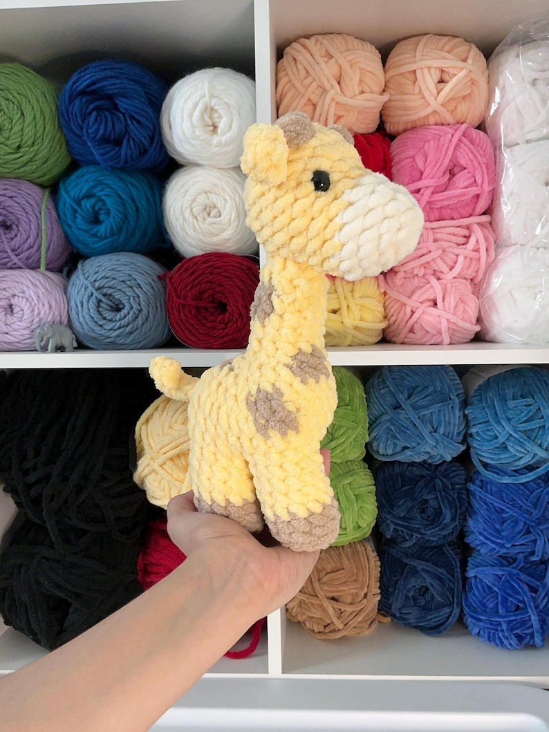 Pu&ograve; includere: Una giraffa all'uncinetto gialla e marrone con macchie marroni. La giraffa &egrave; tenuta davanti a una mensola piena di gomitoli di lana di vari colori, tra cui blu, verde, rosa e bianco. La giraffa &egrave; un giocattolo morbido.