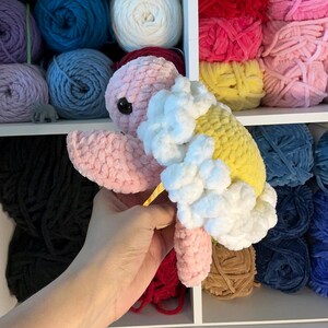 Pu&ograve; includere: Una tartaruga all'uncinetto fatta a mano in rosa, giallo e bianco. La tartaruga ha un occhio nero ed &egrave; tenuta davanti a uno scaffale pieno di gomitoli di filato colorati. Il guscio della tartaruga &egrave; giallo con accenti bianchi soffici.