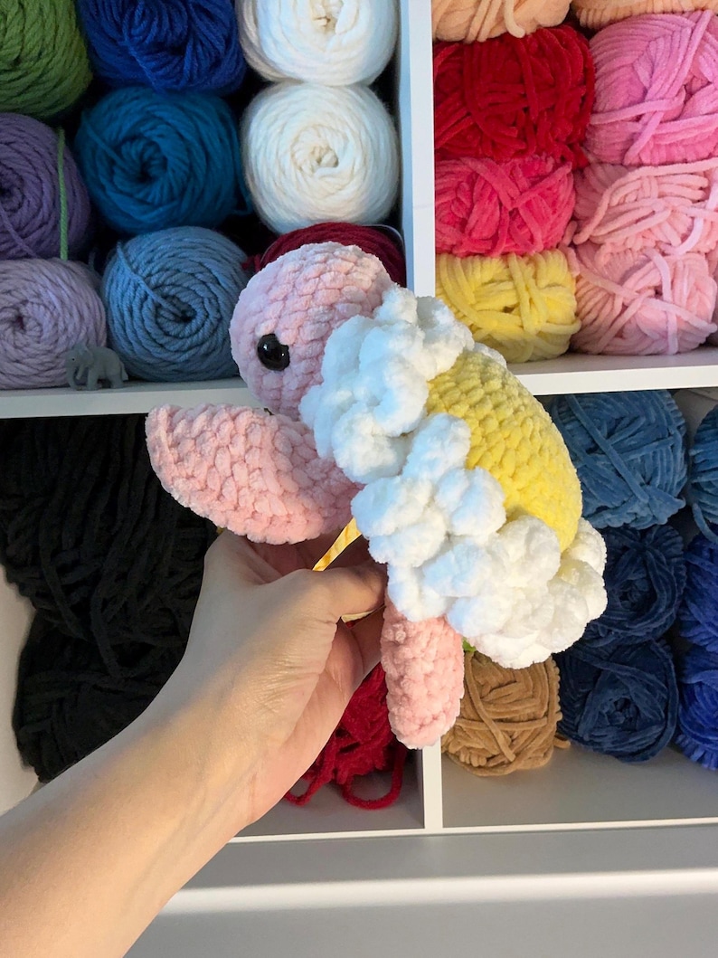 Pu&ograve; includere: Una tartaruga all'uncinetto fatta a mano in rosa, giallo e bianco. La tartaruga ha un occhio nero ed &egrave; tenuta davanti a uno scaffale pieno di gomitoli di filato colorati. Il guscio della tartaruga &egrave; giallo con accenti bianchi soffici.