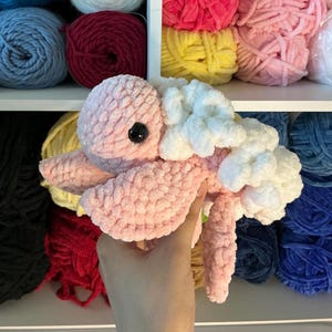 Pu&ograve; includere: Una medusa rosa di peluche fatta a mano, con dettagli bianchi e un occhio nero. La medusa &egrave; realizzata con filato morbido e strutturato. Gli scaffali sullo sfondo sono pieni di gomitoli di filato colorati in vari colori, tra cui rosa, blu e rosso.