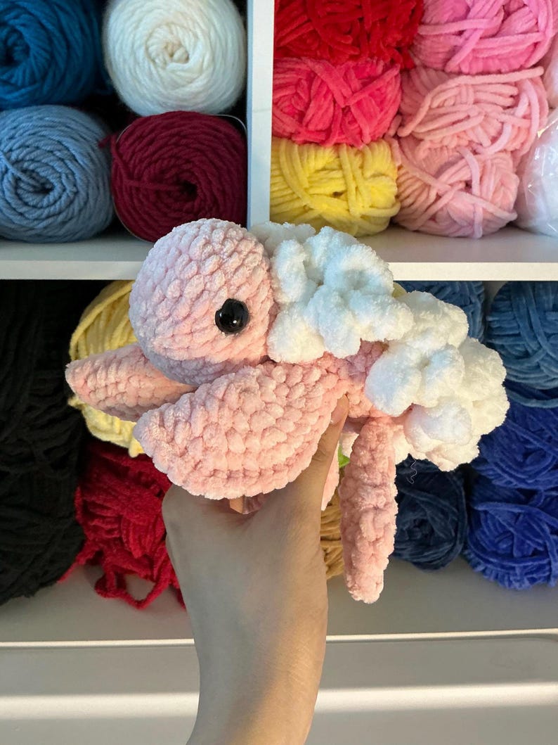 Pu&ograve; includere: Una medusa rosa di peluche fatta a mano, con dettagli bianchi e un occhio nero. La medusa &egrave; realizzata con filato morbido e strutturato. Gli scaffali sullo sfondo sono pieni di gomitoli di filato colorati in vari colori, tra cui rosa, blu e rosso.