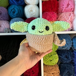 Pu&ograve; includere: Un peluche all'uncinetto di un personaggio con grandi orecchie, una testa verde chiaro e un corpo marrone. Il giocattolo ha grandi occhi neri e un piccolo sorriso. Lo sfondo presenta scaffali pieni di fili di vari colori.