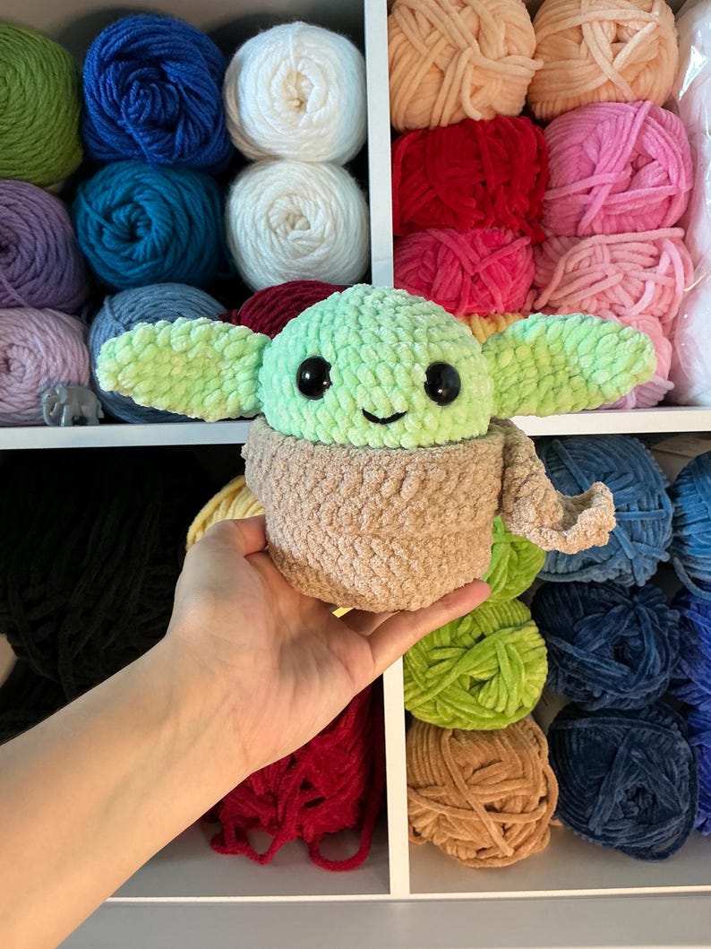 Pu&ograve; includere: Un peluche all'uncinetto di un personaggio con grandi orecchie, una testa verde chiaro e un corpo marrone. Il giocattolo ha grandi occhi neri e un piccolo sorriso. Lo sfondo presenta scaffali pieni di fili di vari colori.