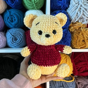 Pu&ograve; includere: Un orsacchiotto di peluche fatto a mano in giallo e bordeaux. L'orso &egrave; fatto di filato morbido e strutturato e ha occhi neri a bottone. L'orso &egrave; tenuto in mano da una persona, con vari gomitoli di filato colorato sullo sfondo.