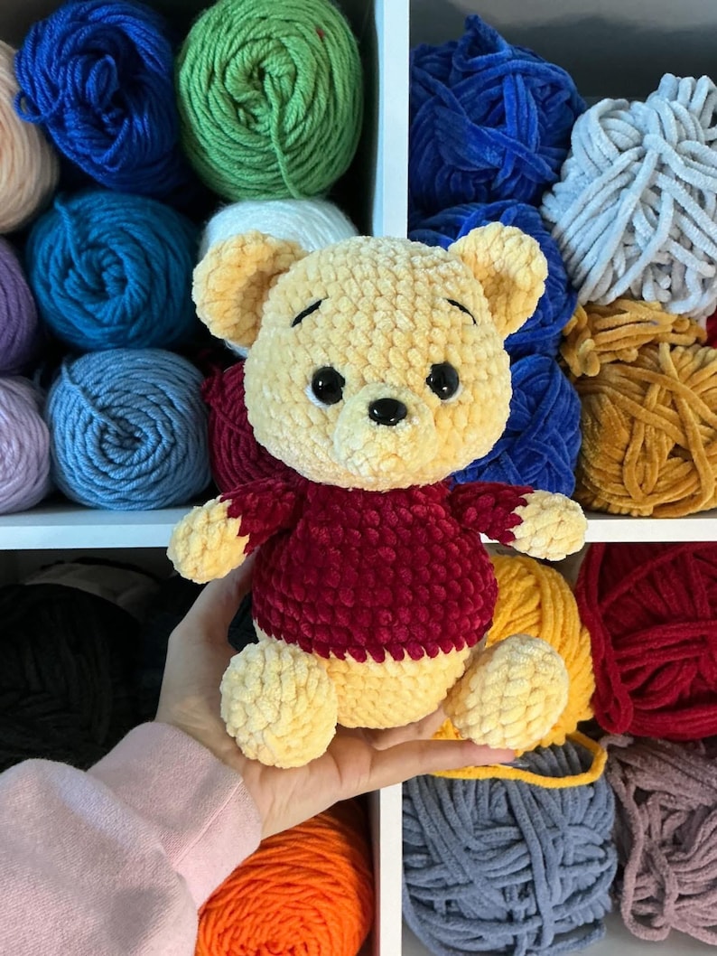Pu&ograve; includere: Un orsacchiotto di peluche fatto a mano in giallo e bordeaux. L'orso &egrave; fatto di filato morbido e strutturato e ha occhi neri a bottone. L'orso &egrave; tenuto in mano da una persona, con vari gomitoli di filato colorato sullo sfondo.