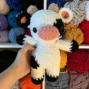Pu&ograve; includere: Un peluche di mucca all'uncinetto bianco e nero fatto a mano con un muso e orecchie interne rosa. La mucca ha occhi neri ed &egrave; circondata da gomitoli di filato colorati su uno scaffale. Il peluche &egrave; morbido e coccoloso.