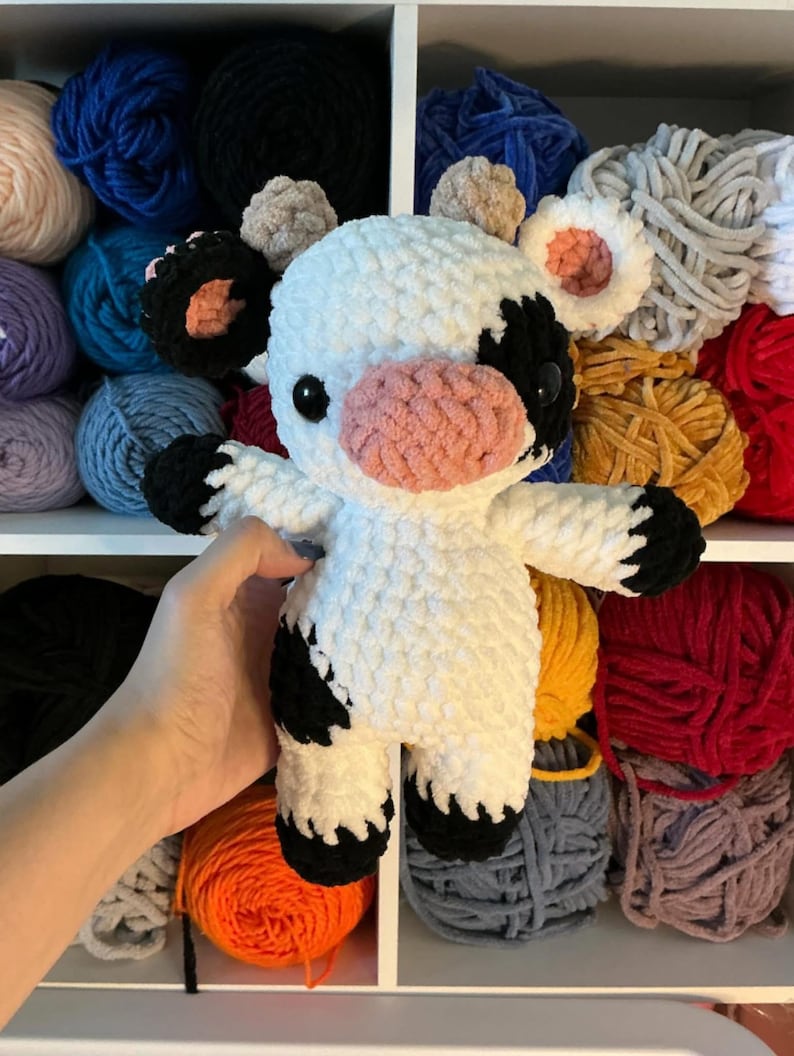 Pu&ograve; includere: Un peluche di mucca all'uncinetto bianco e nero fatto a mano con un muso e orecchie interne rosa. La mucca ha occhi neri ed &egrave; circondata da gomitoli di filato colorati su uno scaffale. Il peluche &egrave; morbido e coccoloso.