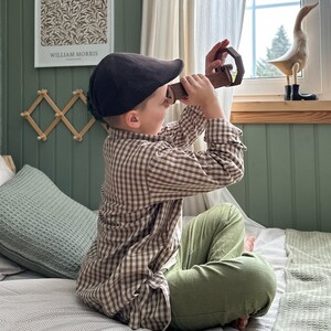 May include: A young boy wearing a brown and white plaid shirt and a brown newsboy cap sits on a bed with a green pillow. He is looking through a wooden toy periscope at a wooden duck figurine on a shelf above a window.