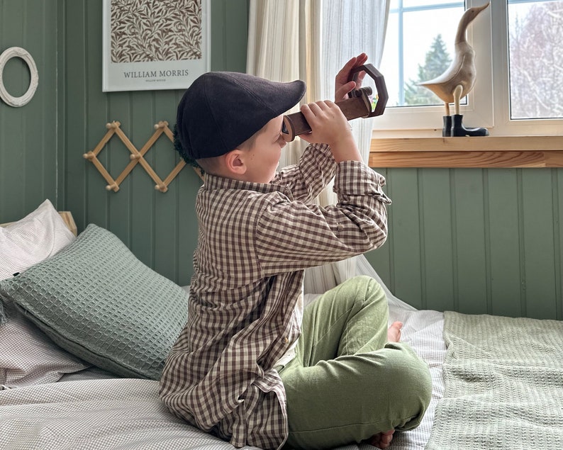 May include: A young boy wearing a brown and white plaid shirt and a brown newsboy cap sits on a bed with a green pillow. He is looking through a wooden toy periscope at a wooden duck figurine on a shelf above a window.