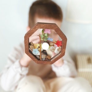 May include: A wooden octagonal frame with a clear glass insert filled with colorful flowers, leaves, and small stones. The frame is held by a child.