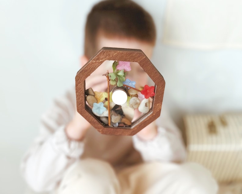 May include: A wooden octagonal frame with a clear glass insert filled with colorful flowers, leaves, and small stones. The frame is held by a child.