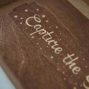 May include: A dark brown wooden box lid featuring the engraved text Capture the in a cursive script, surrounded by small engraved star and dot patterns. The wood grain is visible across the surface, and the lid has a rectangular shape.