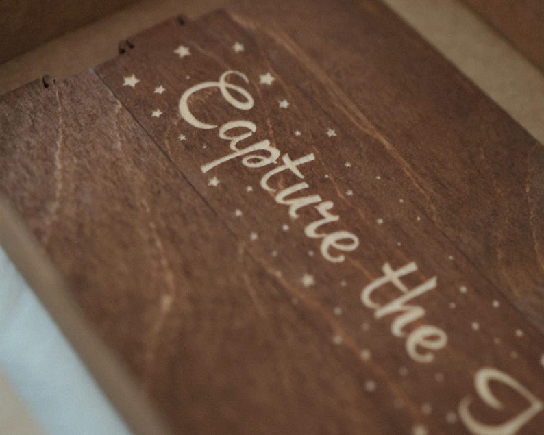 May include: A dark brown wooden box lid featuring the engraved text Capture the in a cursive script, surrounded by small engraved star and dot patterns. The wood grain is visible across the surface, and the lid has a rectangular shape.