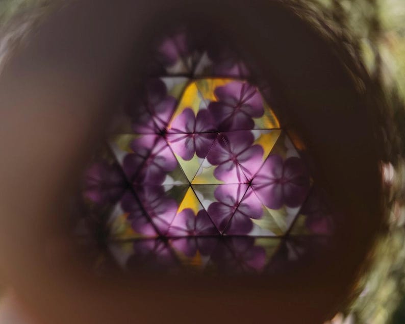 May include: A view through a kaleidoscope showing a repeating geometric pattern of purple flower petals. The central design features a symmetrical arrangement of violet-colored floral shapes reflected against yellow and white triangular segments.