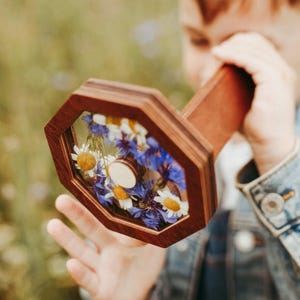 May include: A wooden kaleidoscope with an octagonal viewing end featuring a clear panel filled with pressed blue cornflowers and white daisies with yellow centers. The device has a dark brown wooden frame and a long, cylindrical wooden handle.