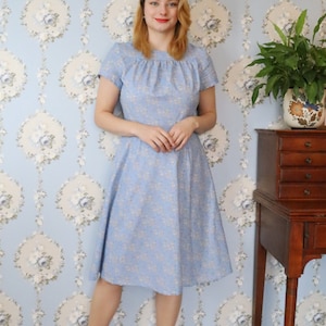 May include: A woman wearing a light blue dress with a floral pattern stands in front of a blue and white floral wallpaper. She is wearing brown shoes and has her hair pulled back.