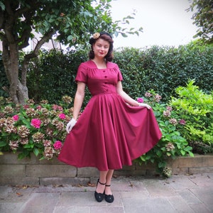 May include: A woman wearing a maroon dress with a white collar and buttons, a flower in her hair, and white gloves. She is standing in a garden with green bushes and pink flowers.