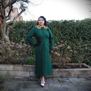 Op de afbeelding: Een vrouw draagt een lange, groene jas met een bruine bontkraag. Ze staat op een betegeld terras voor een groene haag.