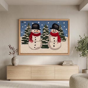May include: A rectangular punch needle wall art piece in a light wood frame featuring two snowmen wearing black top hats and red scarves standing in a snowy landscape with dark green pine trees against a blue sky with white falling snowflakes.