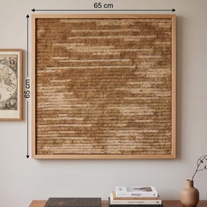 May include: A square wall art piece featuring a textured, horizontal brown fiber design encased in a light wood frame. The artwork measures 65 cm by 65 cm as indicated by the dimension lines. The surface consists of layered, shaggy brown material.