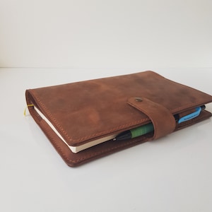 May include: A brown leather notebook cover with visible stitching along the edges. The cover features a central snap closure strap and a side loop holding a green and black pen. A small yellow ribbon bookmark extends from the side of the pages.