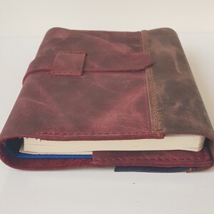 May include: A maroon leather journal cover with a distressed texture and a matching strap closure. The cover features visible stitching along the edges and holds a notebook with cream-colored pages and a blue spine visible at the bottom.