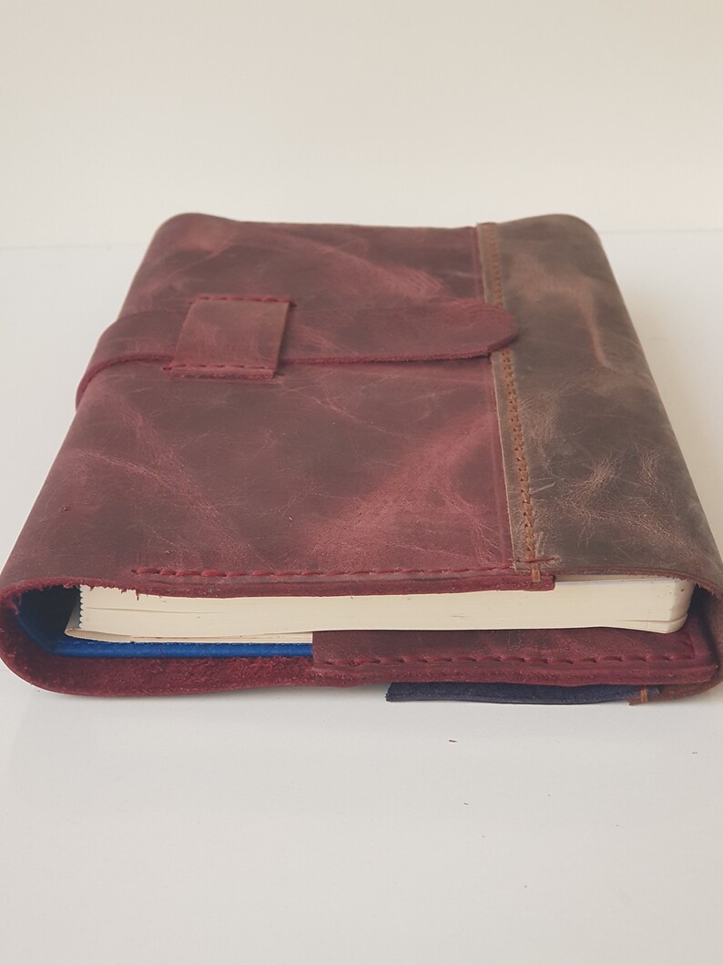 May include: A maroon leather journal cover with a distressed texture and a matching strap closure. The cover features visible stitching along the edges and holds a notebook with cream-colored pages and a blue spine visible at the bottom.