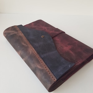 May include: A rectangular leather journal cover featuring a distressed finish in shades of deep burgundy, dark brown, and a central navy blue panel. The cover is secured with a single brass-colored metal snap button and features visible stitching.