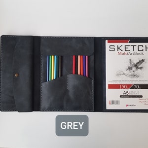 May include: A dark grey leather sketchbook cover featuring a flap with a brass snap closure, a central pocket containing ten colored pencils, and a right-side sleeve holding an A5 Sketch MultiArtBook with 25 sheets of 150g paper.