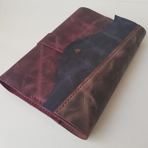 May include: A leather journal cover featuring a tri-color design with panels in deep burgundy, dark navy blue, and brown. The cover has visible stitching along the edges and a single metallic snap button closure on the front flap to secure the contents.