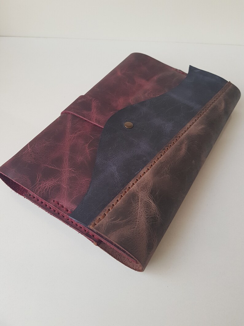 May include: A leather journal cover featuring a tri-color design with panels in deep burgundy, dark navy blue, and brown. The cover has visible stitching along the edges and a single metallic snap button closure on the front flap to secure the contents.