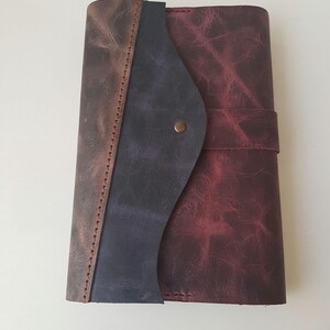 May include: A rectangular leather journal featuring a distressed finish in shades of deep burgundy, dark blue, and brown. The cover has a curved flap closure secured with a single brass-colored metal stud, and visible stitching along the spine.