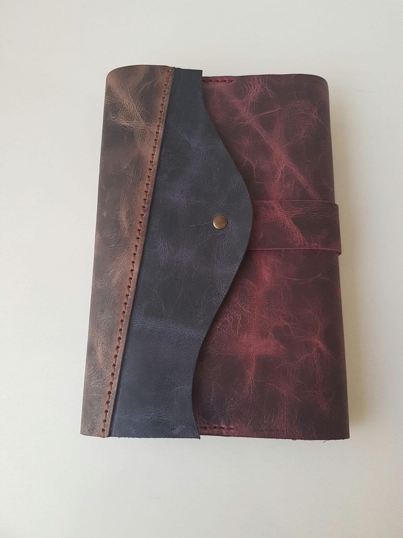 May include: A rectangular leather journal featuring a distressed finish in shades of deep burgundy, dark blue, and brown. The cover has a curved flap closure secured with a single brass-colored metal stud, and visible stitching along the spine.